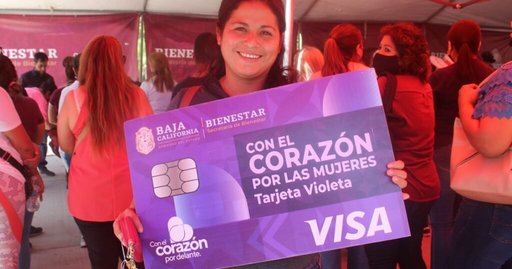 Una mujer sostiene orgullosa una gran tarjeta representativa de la Tarjeta Violeta, un programa de apoyo económico bimestral dirigido a mujeres jefas de familia en Baja California. La imagen muestra un evento de entrega del programa, destacando el compromiso del gobierno estatal en mejorar las condiciones de vida de las mujeres en situación de vulnerabilidad.